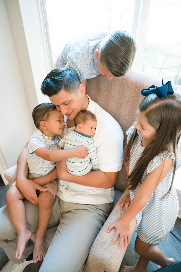 A father holds his newborn son in a rocker while surrounded by his older three children during a lifestyle newborn photoshoot in Houston TX. 