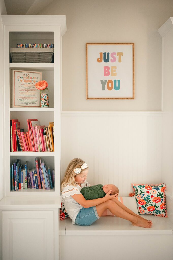 A young girl holds her newborn baby brother next to a shelf of colorful books during an in-home photoshoot with cypress newborn photographer Mel B Photo 