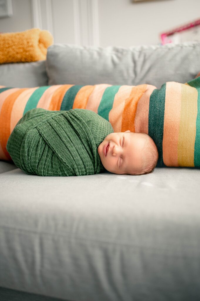 A newborn baby boy smiles in a green swaddle during an in-home session with a cypress Newborn Photographer Tip