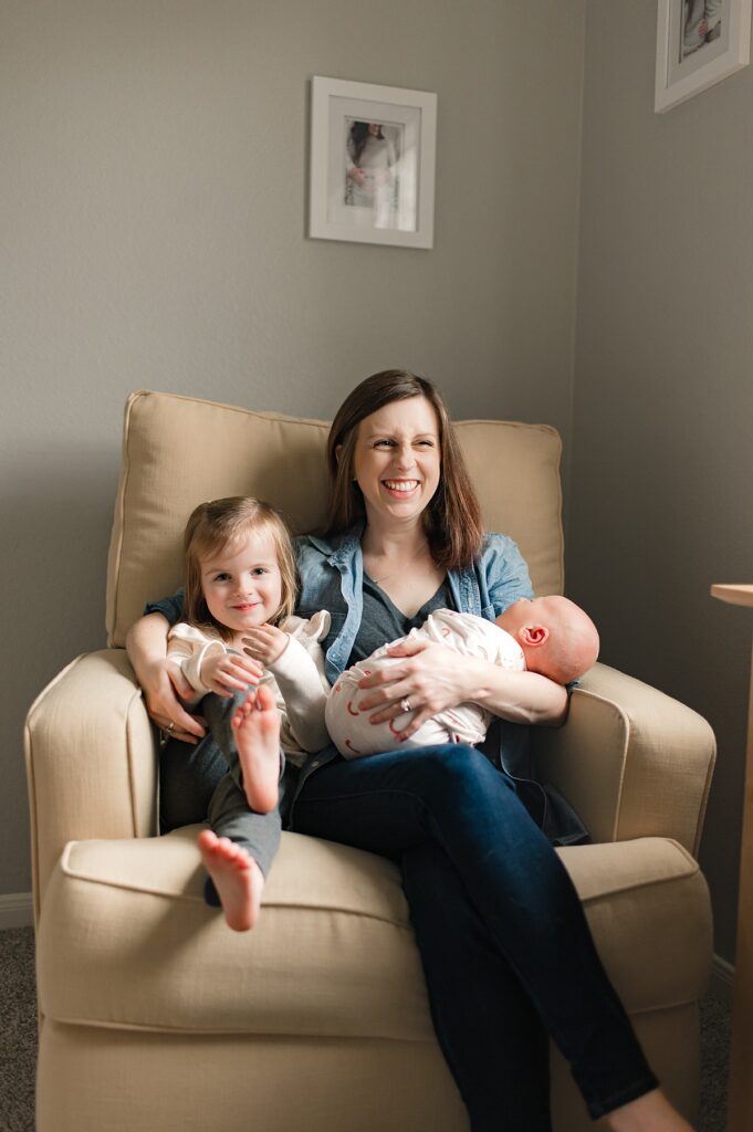 A Texas mother is shown cradling her newborn son in his neutral nursery next to her older daughter during a lifestyle in-home newborn session with Houston area newborn and family photographer, Mel B Photo.