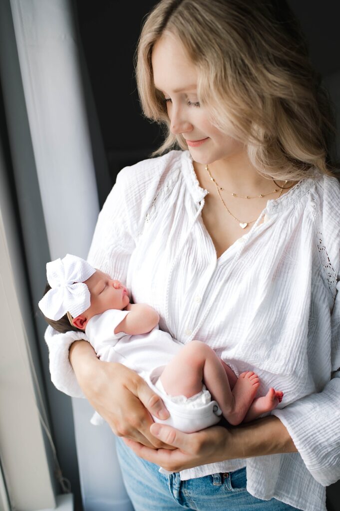 Natural lighting from a window illuminates a Texas mother who is cradling her newborn daughter in her arms during an in-home newborn photoshoot with Houston area photographer, Mel B Photo.
