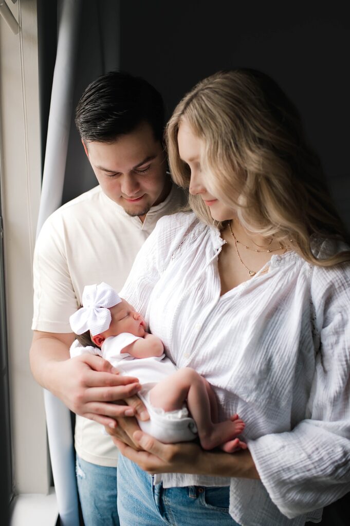 A couple holds their newborn daughter in a white onesie and matching bow during a dark & moody in-home newborn session with Texas newborn photographer, Mel B Photo.