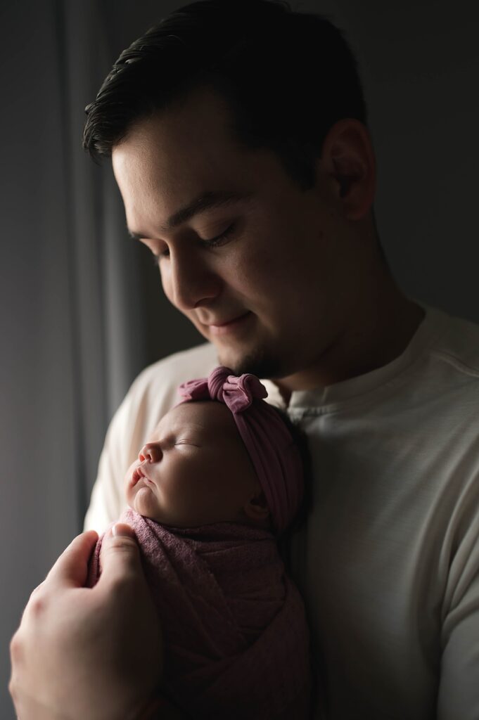 A father holds his newborn daughter in a pink swaddle and matching bow during a dark & moody in-home newborn session with Texas newborn photographer, Mel B Photo.