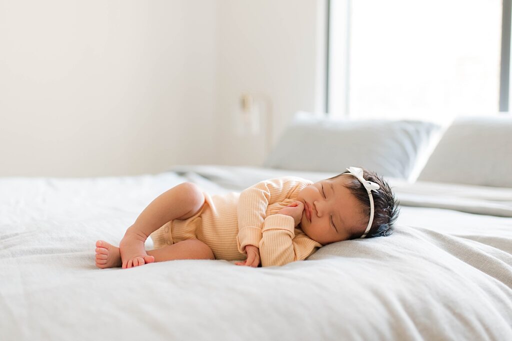 A newborn baby girl wearing a soft yellow sleeper and a white bow in her dark hair sleeps peacefully on a white duvet during her Cypress, TX newborn session with Mel B Photo.