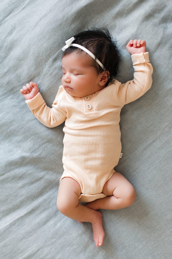 A newborn baby girl wearing a soft yellow sleeper and a white bow in her dark hair sleeps peacefully on her parents bed during her Cypress, TX newborn session with Mel B Photo.