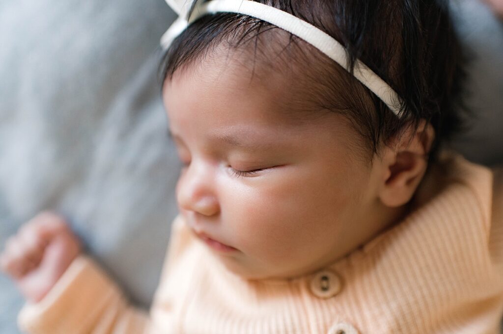 A sleeping newborn baby girl wearing a white bow and a mustard yellow onesie is pictured in a close up during an in-home lifestyle photo session.