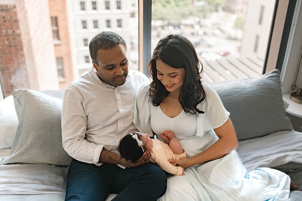 A young couple admire their newborn baby girl lying on her mother's lap as they sit on their gray sofa near large windows with a view of their Texas neighborhood.