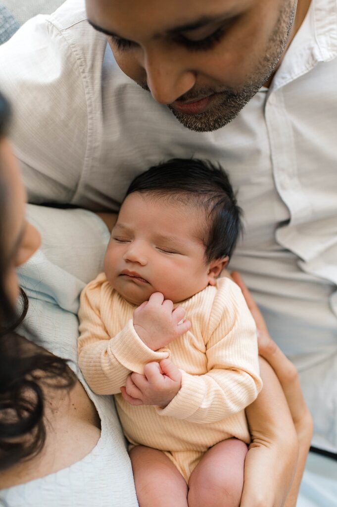 A photo from an in-home lifestyle photo session shows a newborn baby girl with dark hair sleeping in her mother's arms wearing a soft yellow sleeper.