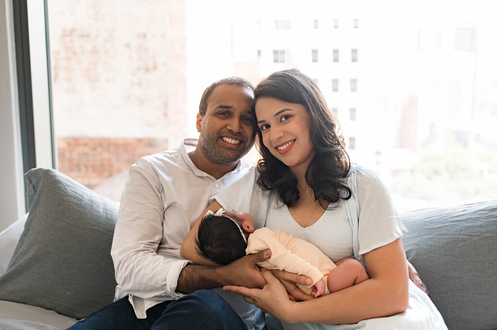 A Texas couple smiles as they hold their newborn daughter with dark hair in their arms seated atop their bed in their naturally lit home.