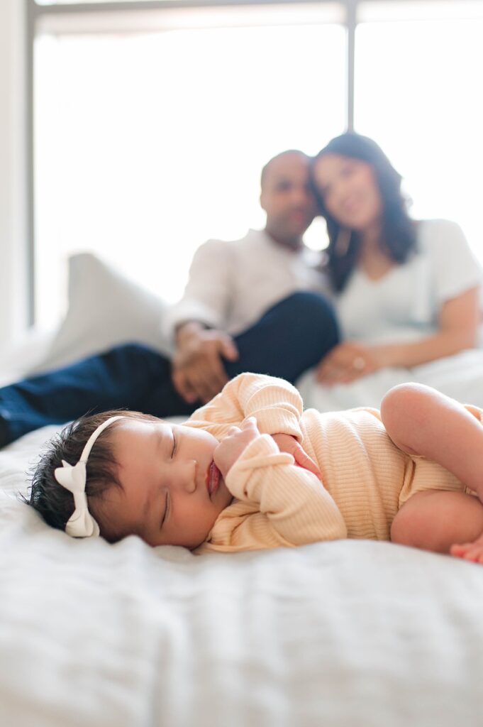 In this photo from an in-home newborn session, a Cypress, TX couple sits before a window in their home on a bed covered in a white linen comforter, as their newborn daughter wearing a yellow onesie and a white bow sleeps before them.