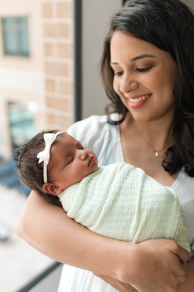 A new mother smiles down upon her swaddled newborn baby girl wearing a small, white bow in her dark hair during their in-home lifestyle photos with Mel B Photo.