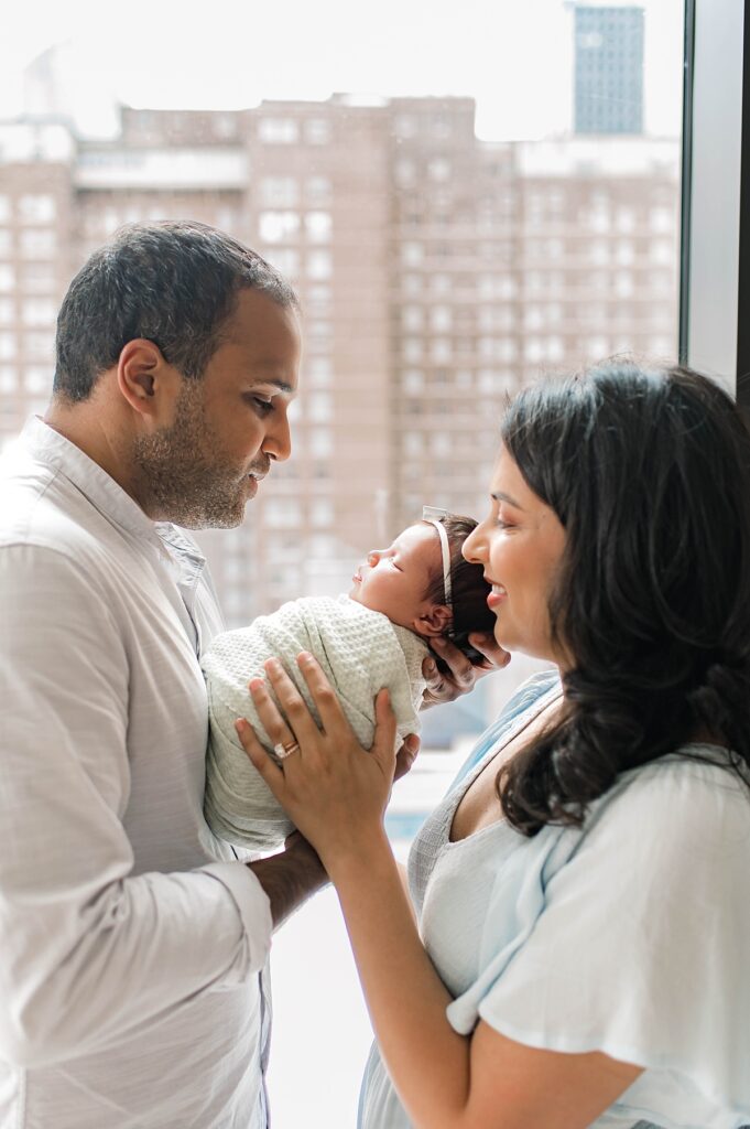 A Cypress, Texas couple are pictured from above holding their newborn daughter wearing a soft yellow onesie near floor-to-ceiling windows during an in-home photo session with Towne Lake newborn photographer, Mel B Photo.