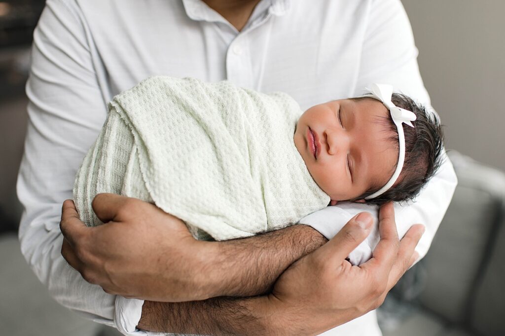 A newborn baby girl swaddled in white muslin wearing a white bow in her dark hair is photographed in her father's arms during an in-home lifestyle photo shoot with Mel B Photo.