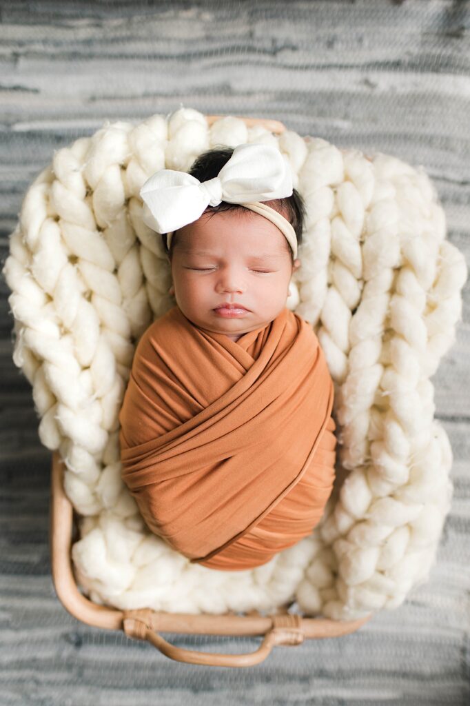 A Cypress, TX newborn is pictured in a burnt orange swaddle atop a chunky knit blanket in a woven Moses basket during an in-home newborn session with Towne Lake newborn photographer, Mel B Photo.