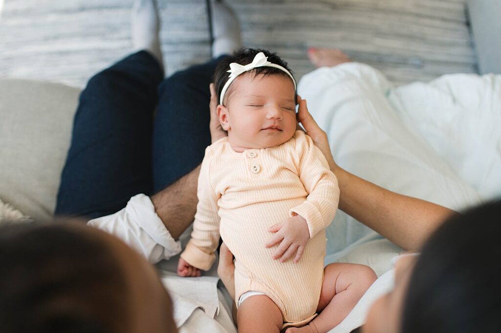 A photo from an in-home lifestyle photo session shows a newborn baby girl with dark hair smiling in her sleep in her mother's arms wearing a soft yellow sleeper.