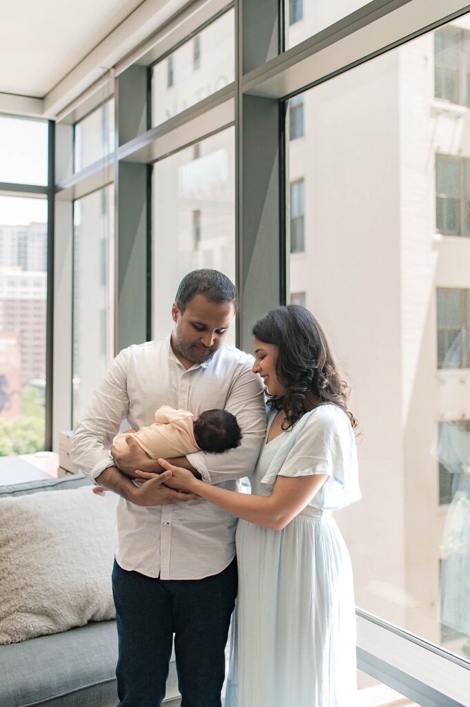 A Cypress, Texas couple are pictured from above holding their newborn daughter wearing a soft yellow onesie near floor-to-ceiling windows during an in-home photo session with Towne Lake newborn photographer, Mel B Photo.