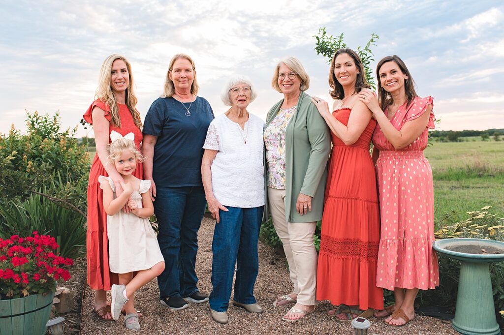 The women of a Tomball, TX family embrace their matriarchs during an outdoor family photo session with Mel B Photo under a blue sky on a gravel patio. 