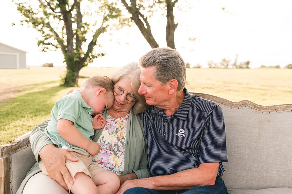 A toddler boy rests his head on his grandmother's chest as she embraces him, seated next to his grandfather during a Texas family photoshoot at golden hour. 