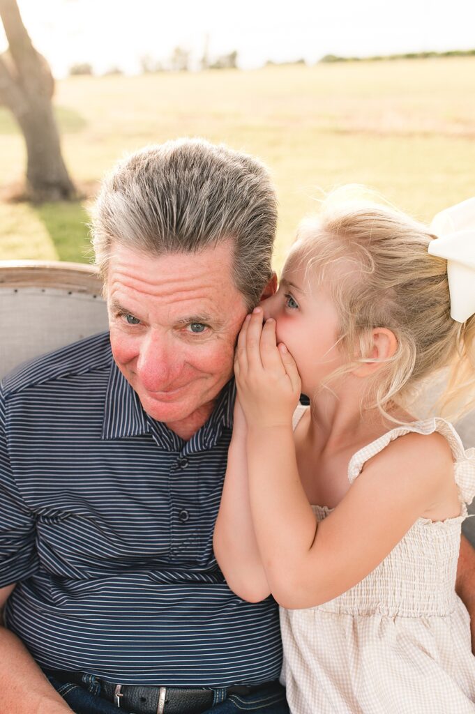 A young blonde girl wearing a white hair bow whispers into her grandfather's ear during a Texas family photoshoot.