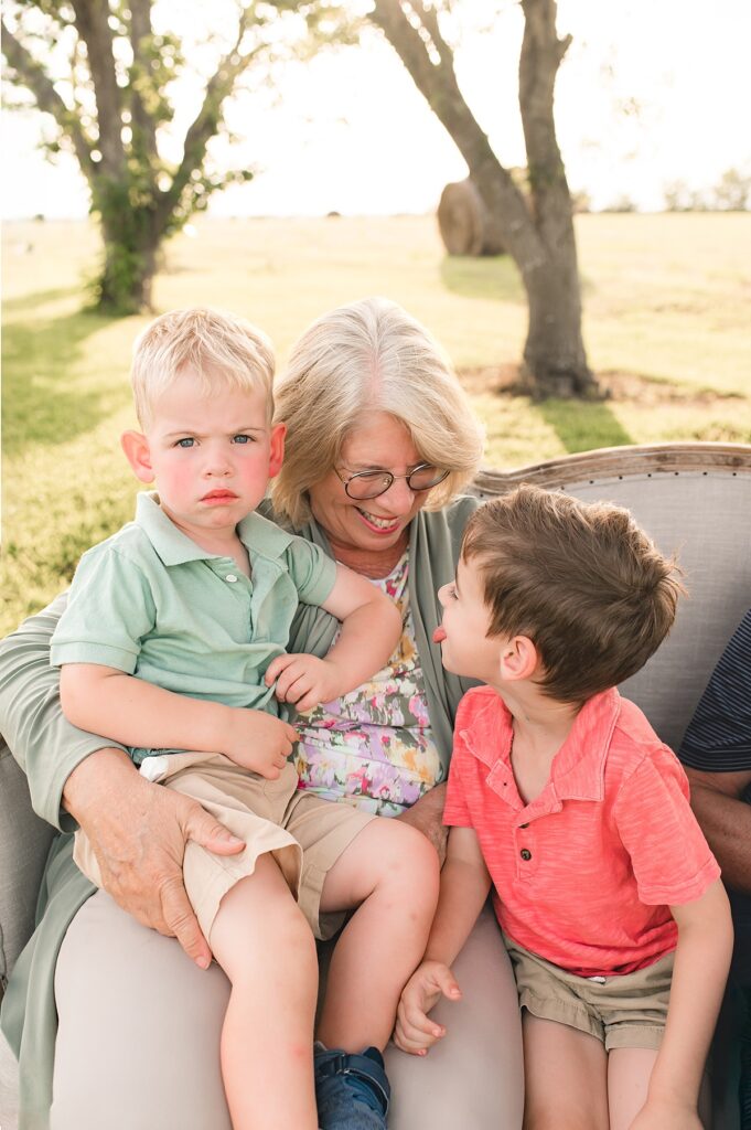 A Tomball, TX grandmother smiles down at her grandson seated next to her on a tan sofa as she holds another grandson on her lap.