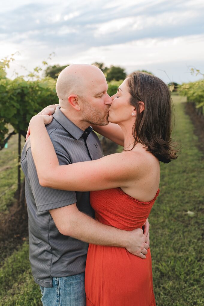 A brunette woman wearing a strapless, red dress kisses her husband during an outdoor family photoshoot with Cypress, TX photographer, Mel B.