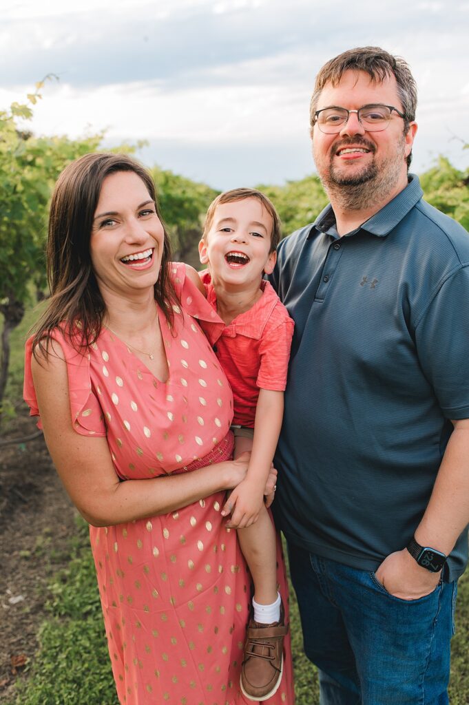 A Texas mother wearing a coral dress smiles next to her husband as she holds her toddler son on her hip during a family photo session with Mel B Photo of Cypress, TX.