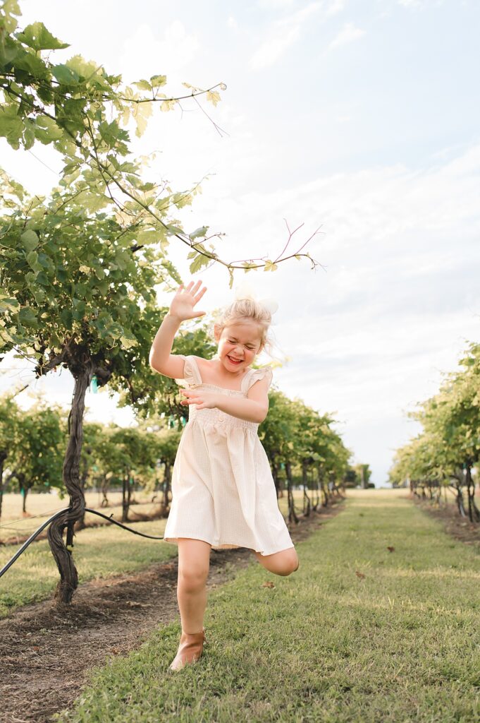 A young blonde girl wearing a beige dress and a white bow runs & laughs with her cousin down a row of grapevines during a Texas family photoshoot.