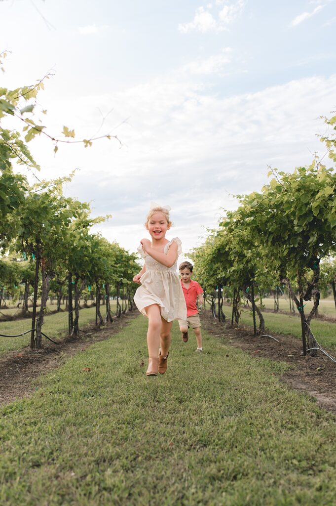 A young blonde girl wearing a beige dress and a white bow runs & laughs with her cousin down a row of grapevines during a Texas family photoshoot.