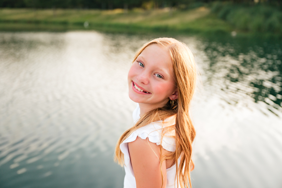 A young girl smiles next to a lake at a Cypress TX park in a photo taken during a family photoshoot with a Cypress photographer.