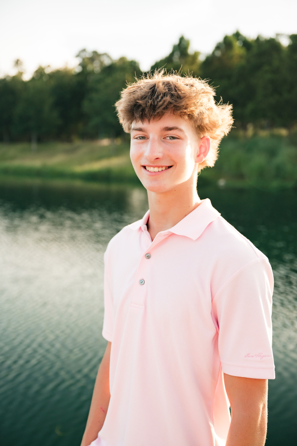 A teen boy in a pink polo shirt smiles next to a Cypress lake in a photo taken by a Cypress photographer for families.