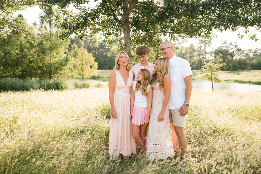 A candid family photo shows parents laughing with their three children beneath the shade of a tree during a family photoshoot with a photographer cypress tx.