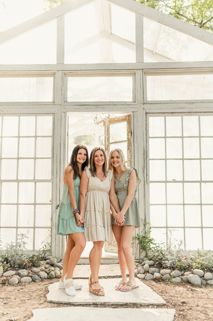 Three ladies of varying ages stand before floor to ceiling paned windows as natural sunlight illuminates from behind them wearing knee length dresses as part of their Conroe studio family photos with Mel B Photo.