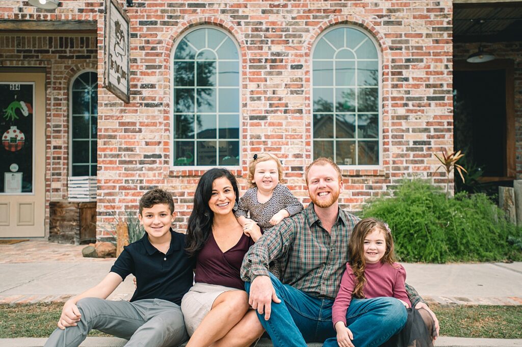 A Tomball family of 5 is pictured seated on a sidewalk before a brick store front during an outdoor family photo session. 