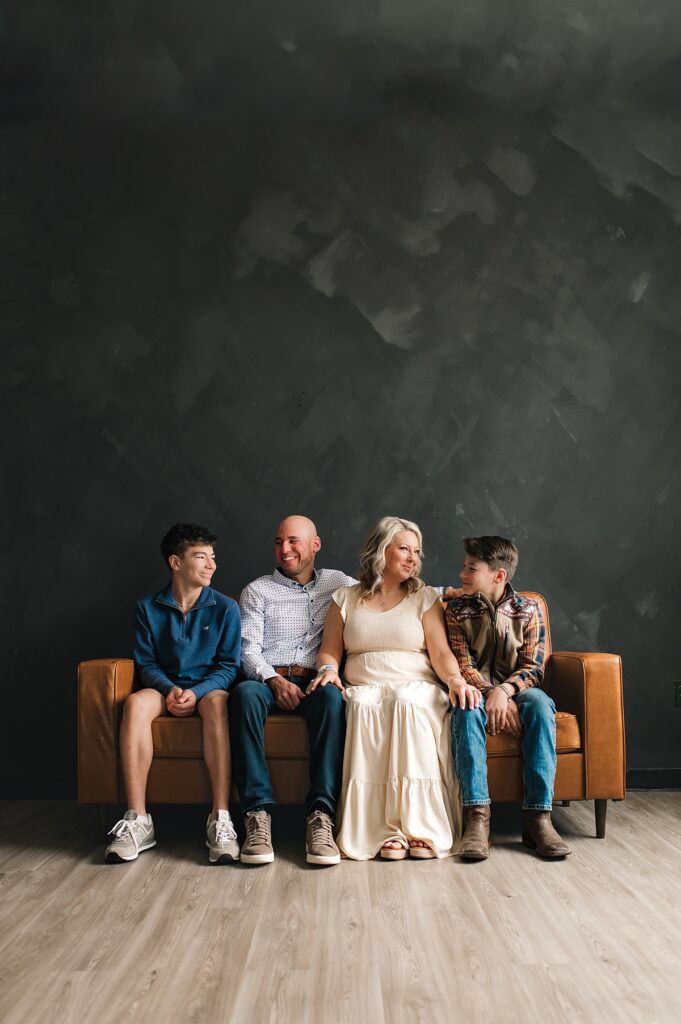 A Tomball family of four is seated on a warm toned leather couch before a black wall for dark & moody Houston family photos. 