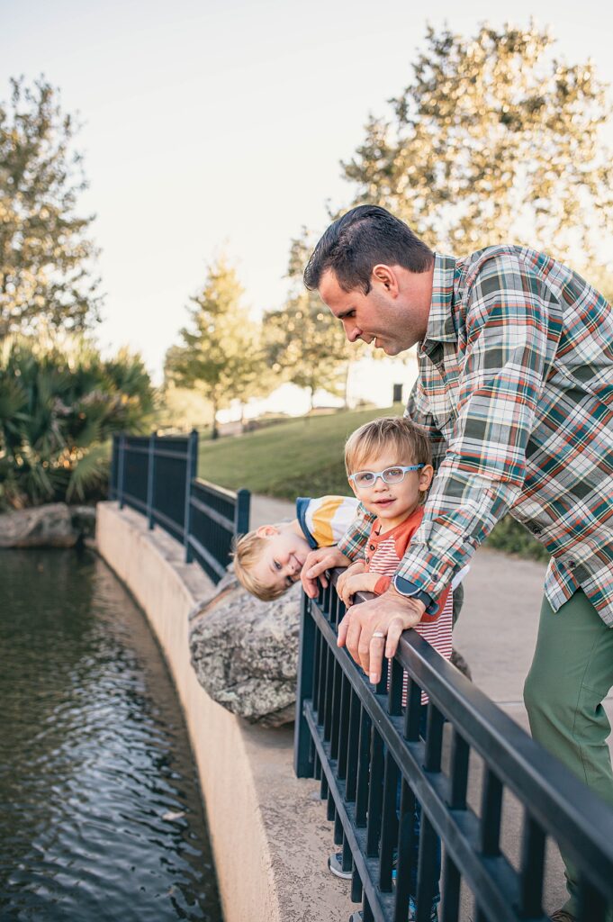 Two young boys lean over the railing at a Cypress park, smiling as their father wearing plaid watches next to them.