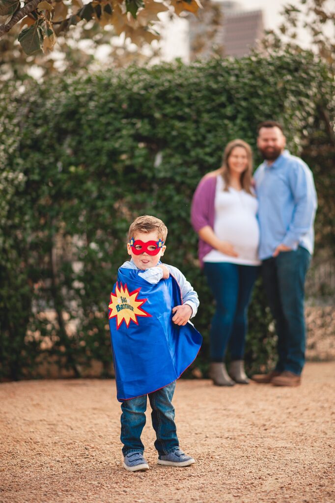 An expectant mother and father stand, out of focus, near shrubs as their toddler son shows off his blue superhero cape that reads 'big brother' during their downtown Houston family photos with Mel B Photo.