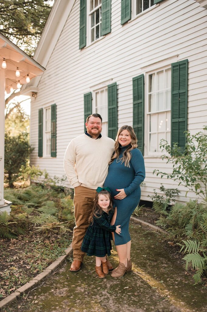 An expectant mother wearing a teal dress embraces her belly as her toddler daughter smiles and hugs her mother's leg as dad, wearing a cream sweater, embraces his wife before a quaint home with green shutters in the Cypress TX area.