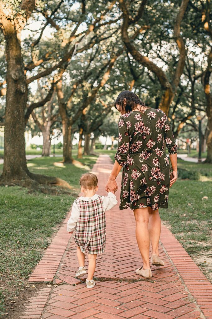 A mother wearing a green floral dress holds her toddler's hand and looks down at ther as they walk away from the photographer along a paved path in a Houston area park. 