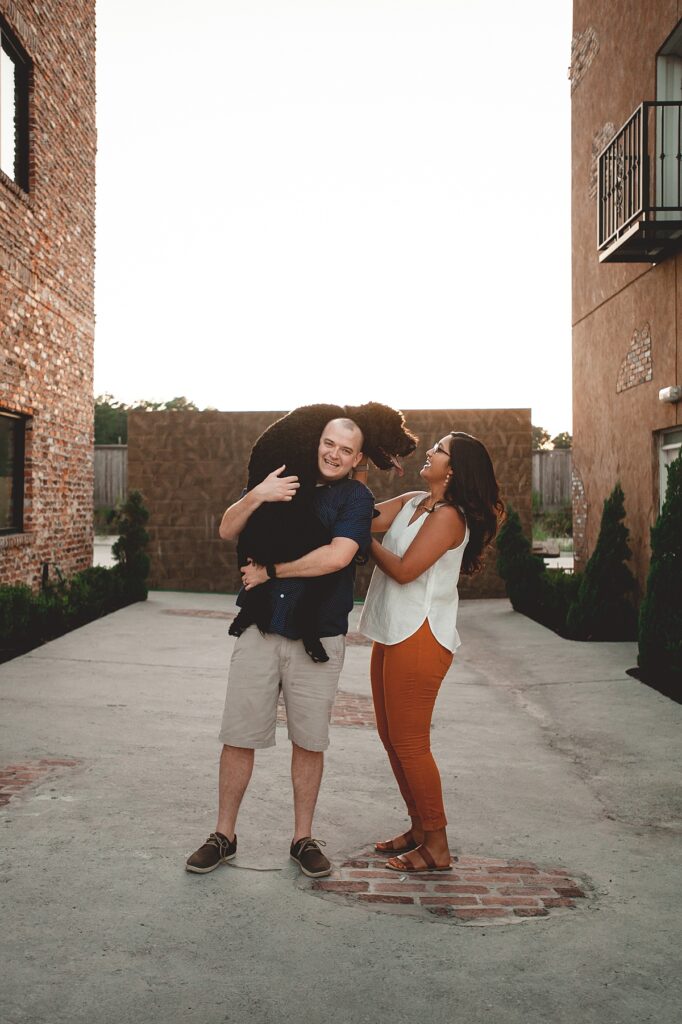 A Texas couple laughs during a family photoshoot with their labradoodle nestled between brick buildings in the downtown Spring region of Houston.