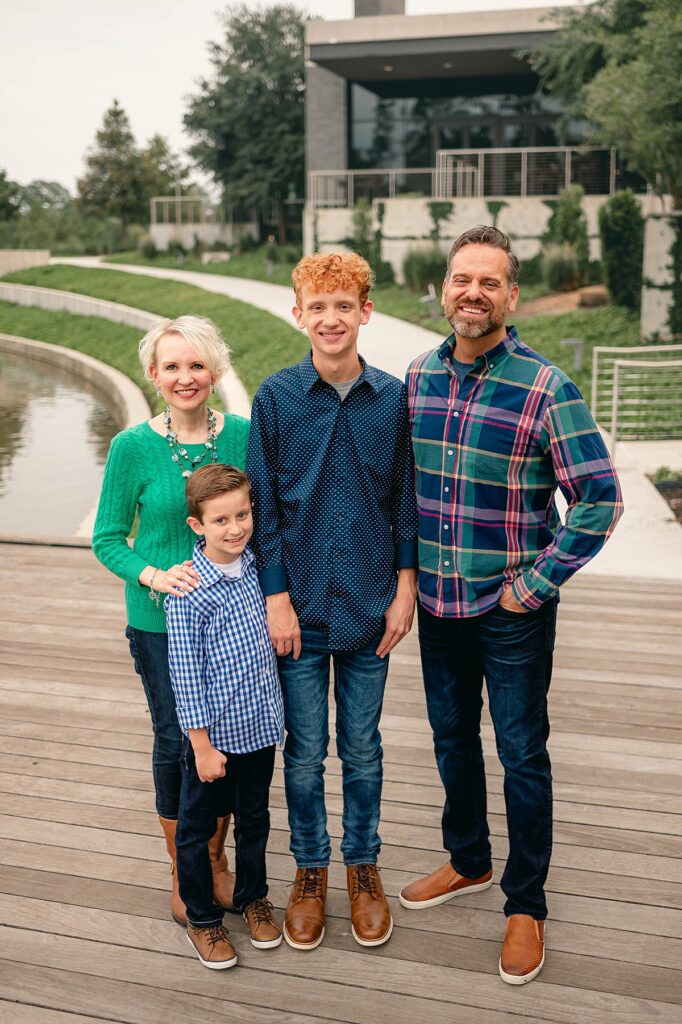 A family of four, wearing jeans and button down shirts pose together before a water feature in the Spring area of Houston, TX for family photos with Mel B Photo.