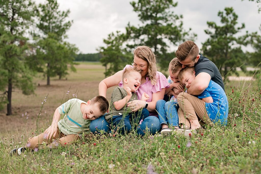 A mother wearing a pink flutter sleeve shirt and a father wearing a deep shade of blue embrace their four laughing sons in a wildflower field in Tomball.