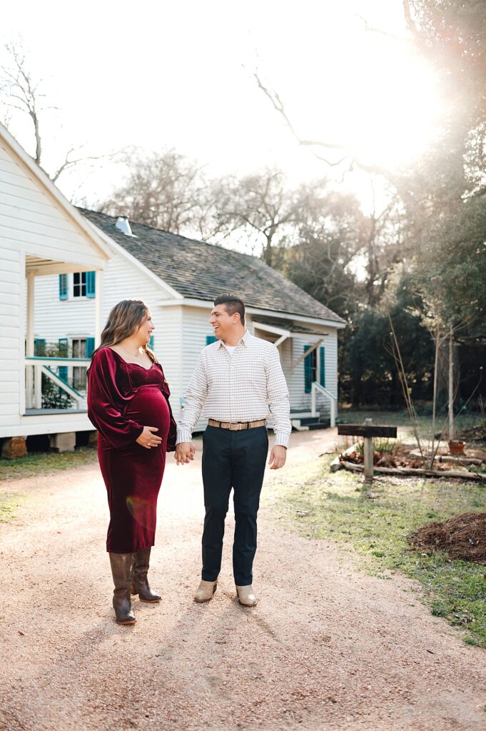 An expectant mother wearing a maroon velvet dress places her right hand on her belly and holds hands with her husband before a public quaint home in the Tomball TX area.