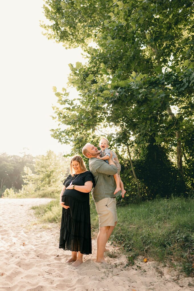 An expectant Texas mother wearing an ankle length black dress on a sandy shore places her hands on the top and bottom of her baby belly as her husband stands next to her, holding their laughing toddler in Spring, TX.