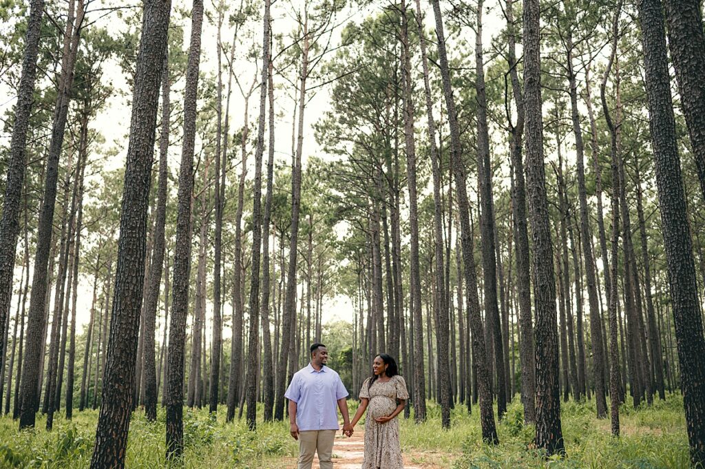 An expectant mother holds hands with her partner as they exchange smiles amongst the trees in The Woodlands during their session with Mel B Phot, Woodlands area family and maternity photographer.