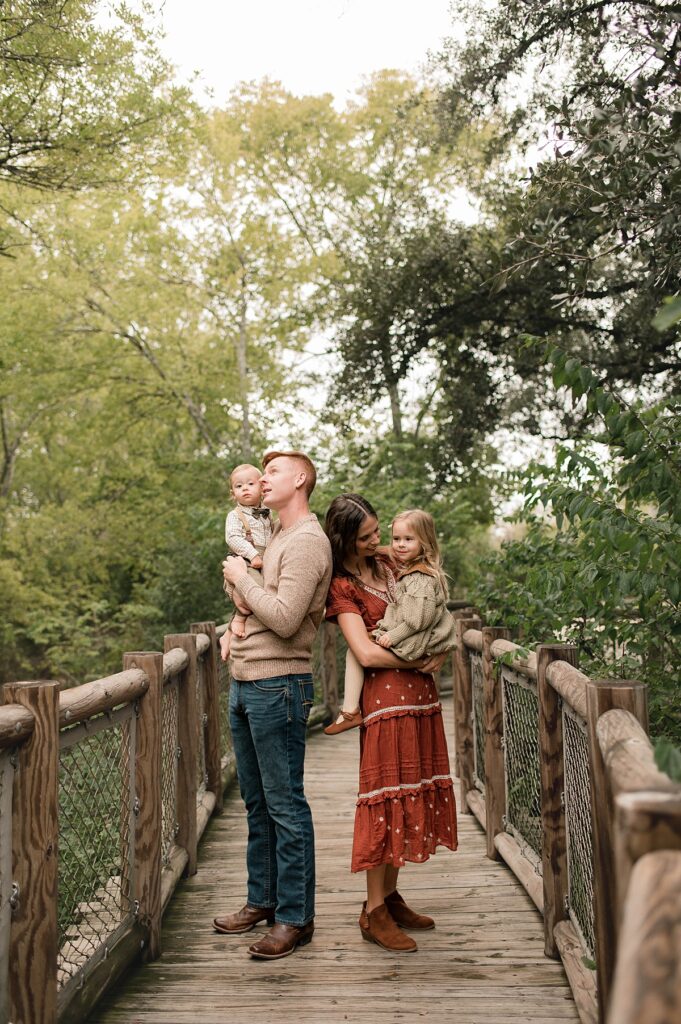 A Texas couple hold their two children on a wooden bridge at a Cypress park surrounded by trees during their family photo session.