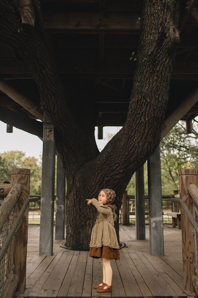 A young girl wearing mary janes and a brown dress points to something in the distance as she explores a Cypress park.