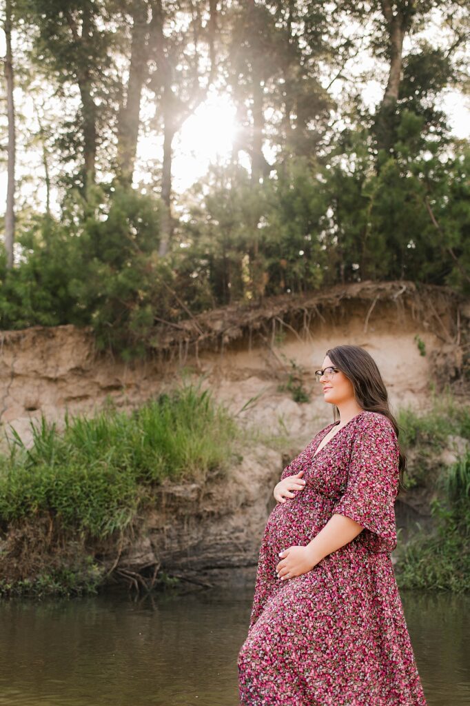 An expectant mother wearing a flowy, maroon floral dress embraces her baby bump as she wades through the water during her Spring maternity session with Mel B Photo.