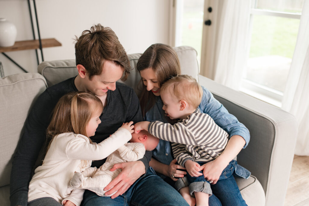 A Houston area family of 5 is shown seated on their greige living room sofa while admiring their newborn as an example of how to plan family photos wardrobe by Mel B Photo.