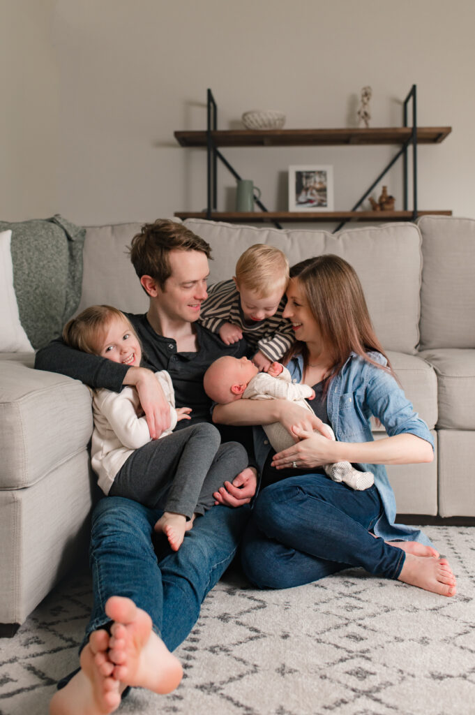 A Tomball TX family photoshoot is depicted as a family of five is seated on the floor of their Houston area home, admiring their newborn baby while wearing coordinating shades of charcoal gray, tan, and denim. 