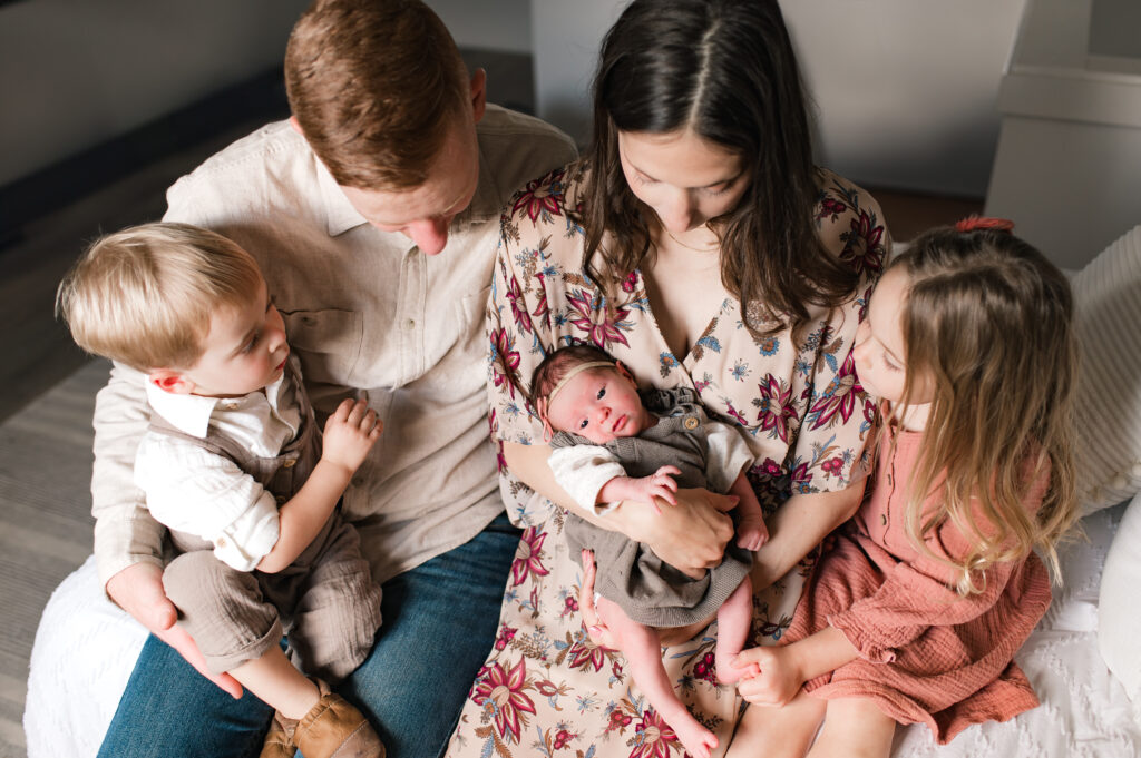 Two young Cypress TX parents look lovingly at their newborn daughter cradled in her mother's arms as their older two children, seated on their parents' laps, admire their baby sister during a family photoshoot with Mel B Photo.